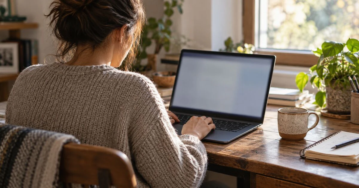 A person reviews a laptop at a sunlit home desk.